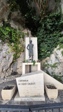 Monumento ai caduti con statue e fiori, situato in una zona rocciosa e verde.