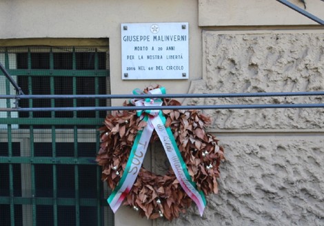 Lapide commemorativa di Giuseppe Malinverni, con una corona di foglie e nastri, apposta su un muro.
