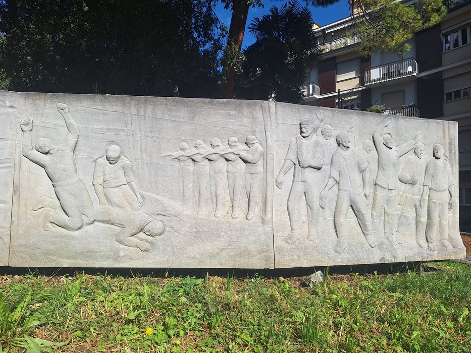 Bassorilievo in pietra con scene di prigionia e deportazione, collocato all'aperto tra alberi e edifici.