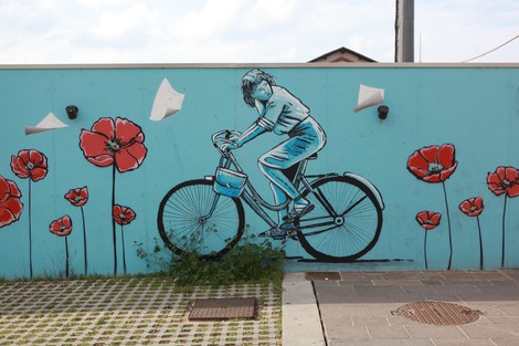 Murales colorato di una ragazza in bicicletta, circondata da papaveri rossi su sfondo azzurro.
