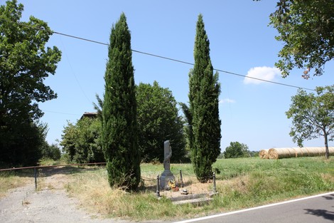 Cippo commemorativo circondato da alti cipressi, in un paesaggio rurale con fieno nel fondo.