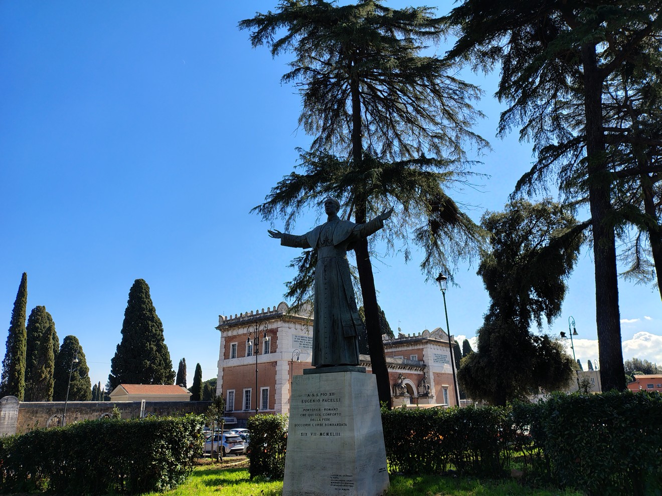Statua di Pacelli in un giardino, con alberi verdi e cielo azzurro sullo sfondo.