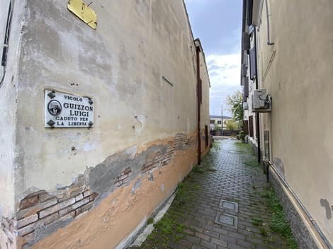 Vicolo stretto con una lapide commemorativa dedicata a Guizzon, circondato da edifici storici.