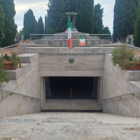 Ingresso monumentale al sacrario, scalinata verso un passaggio sotterraneo con bandiere e iscrizione sopra l'arco.