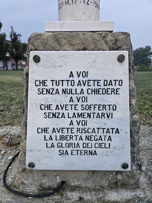 Lapide in marmo con iscrizione, fissata su un cippo in pietra all'aperto, su prato.