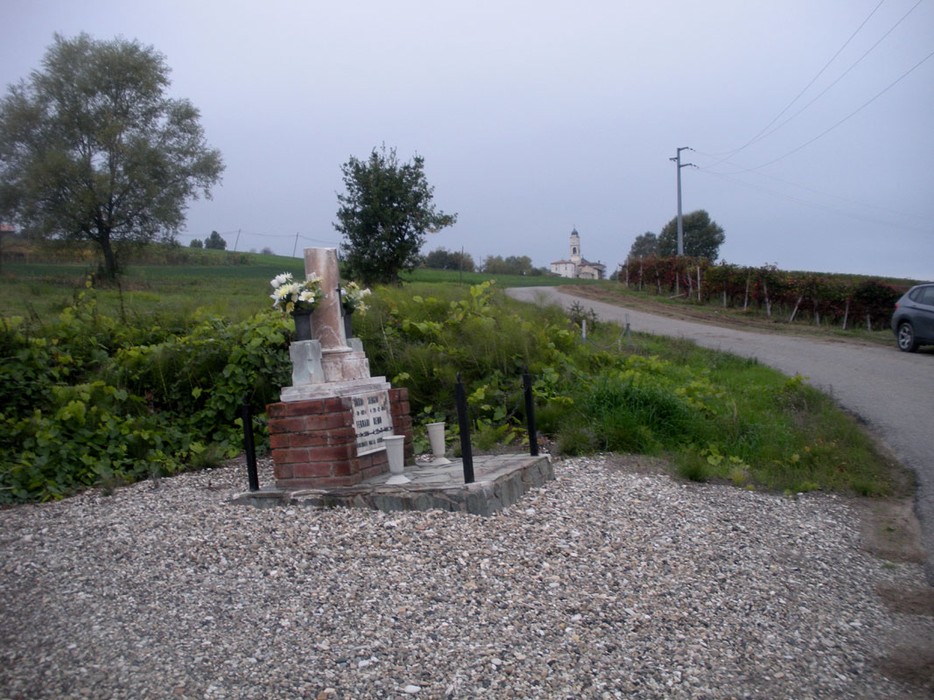 Cippo commemorativo circondato da una verde vegetazione, con una strada che si snoda nelle vicinanze.