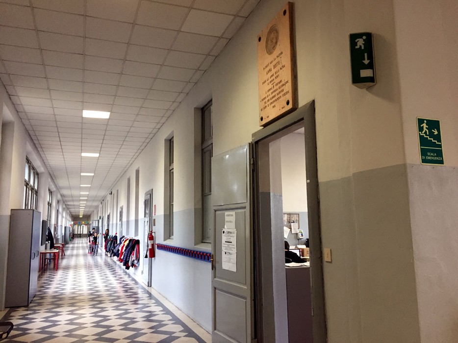 Lapide commemorativa situata in un corridoio scolastico, con giubbotti appesi e segnali di sicurezza.