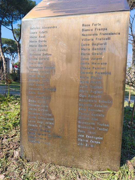 Monumento alle donne cadute per Roma, con una lista di nomi incisi su una placca dorata in un contesto verde.
