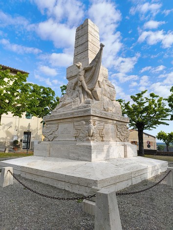 Monumento ai caduti di Piazze, con statua centrale e base monumentale, circondato da alberi e cielo azzurro.