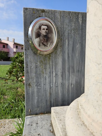 Cippo commemorativo con ritratto di Ricci, nel contesto di un paesaggio verde e case sullo sfondo.