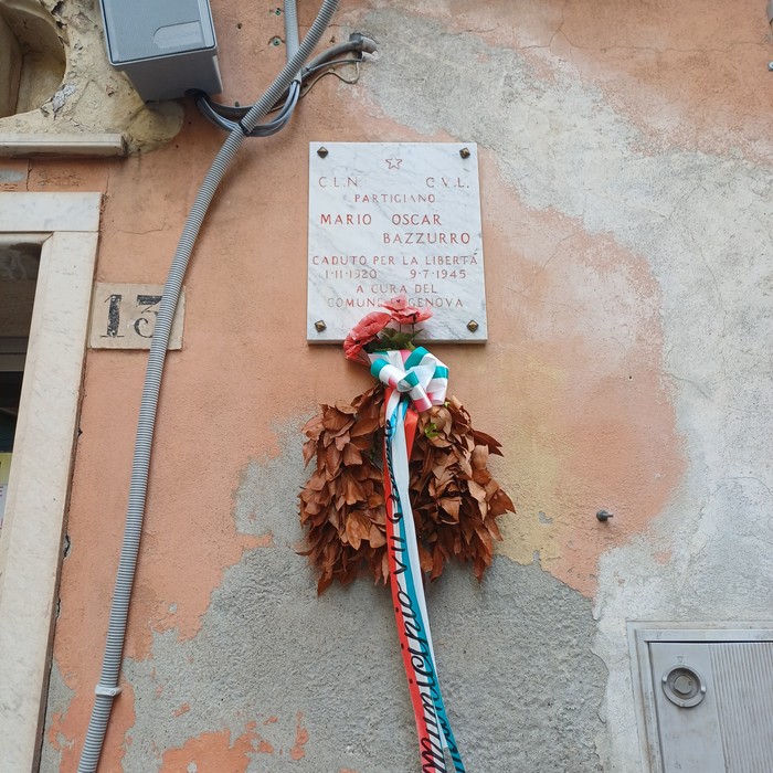 Lapide in marmo su facciata esterna, con iscrizione, corona di foglie e fiori sotto.