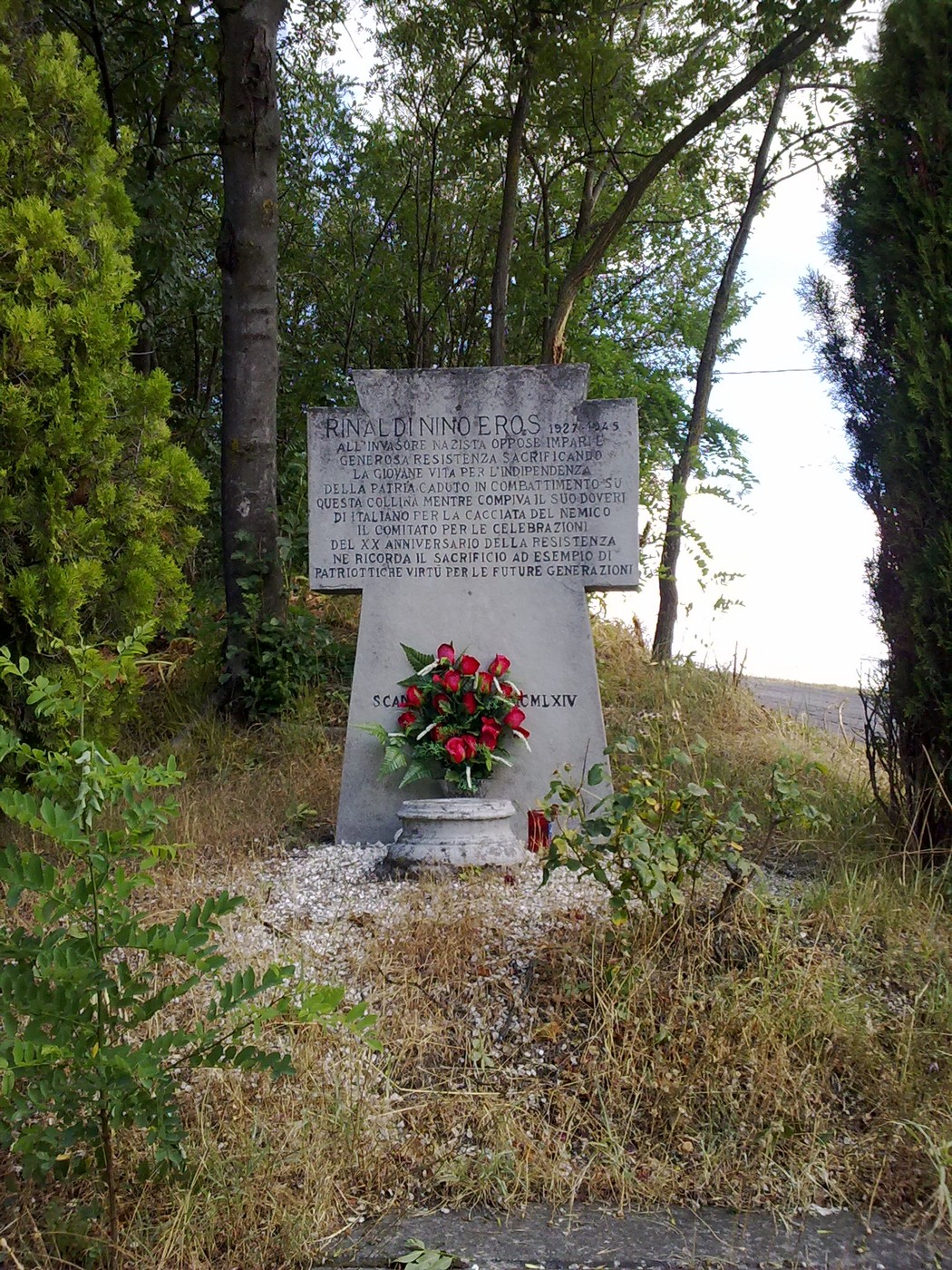 Monumento commemorativo a Rinaldi, circondato da vegetazione, con un omaggio floreale in primo piano.
