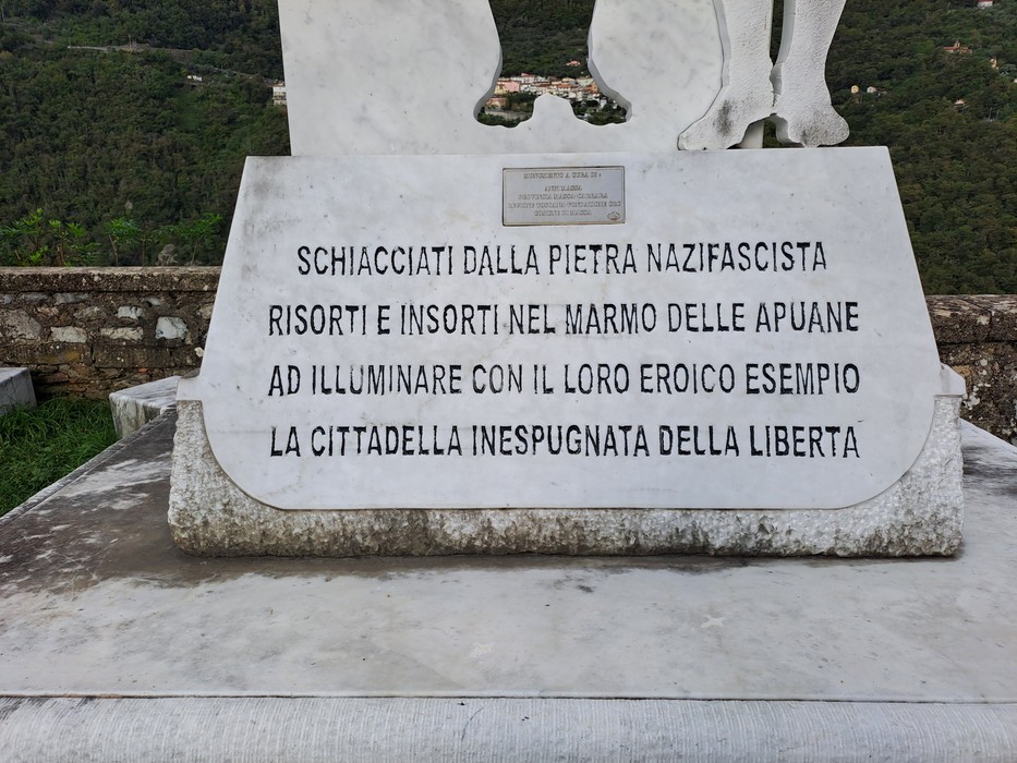 Monumento in marmo bianco con iscrizione, collocato su basamento all'aperto tra il verde.