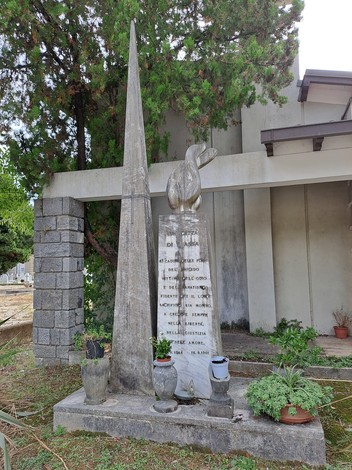 Monumento in pietra con guglia e statua di aquila, lastra con iscrizione, tra alberi presso un edificio.