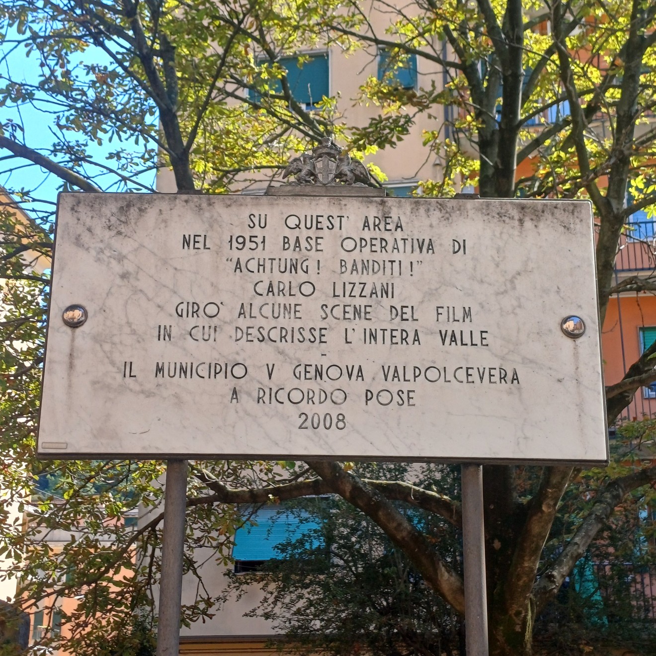 Lapide in marmo con iscrizione, sorretta da due pali metallici tra gli alberi.