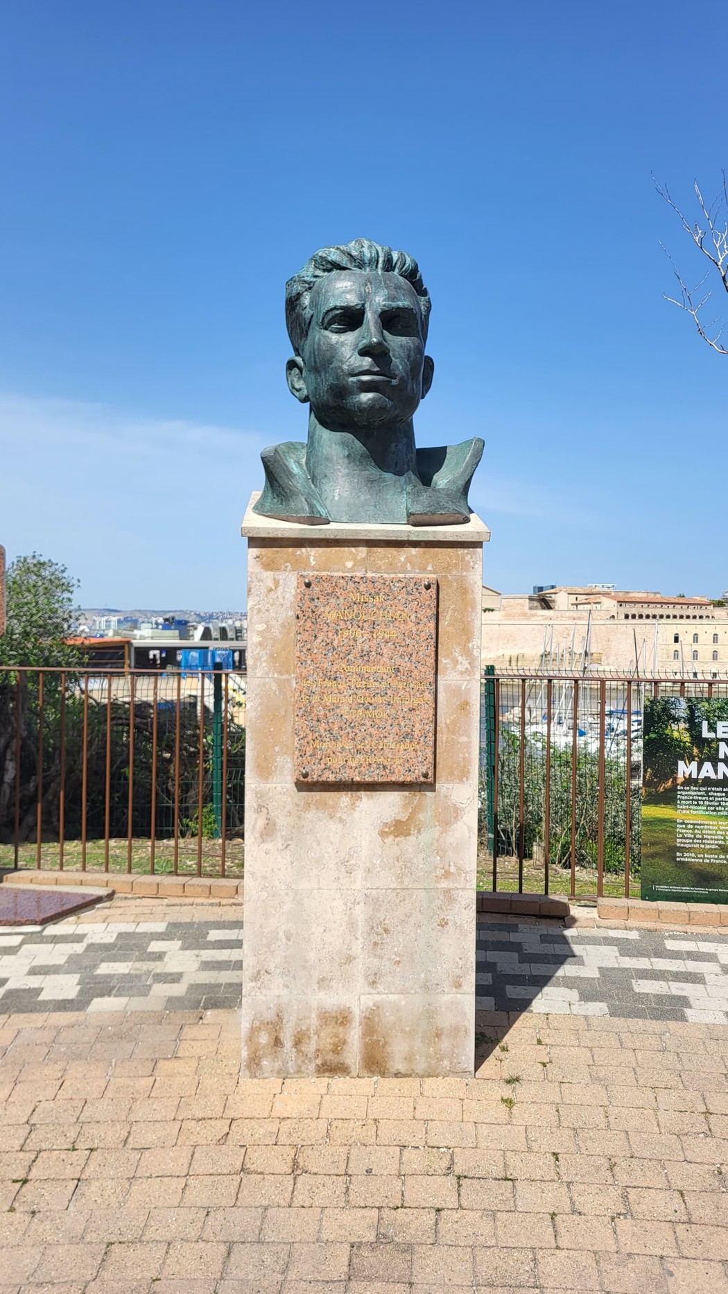 Scultura commemorativa di Missak Manouchian in un giardino, con vista sul paesaggio urbano.