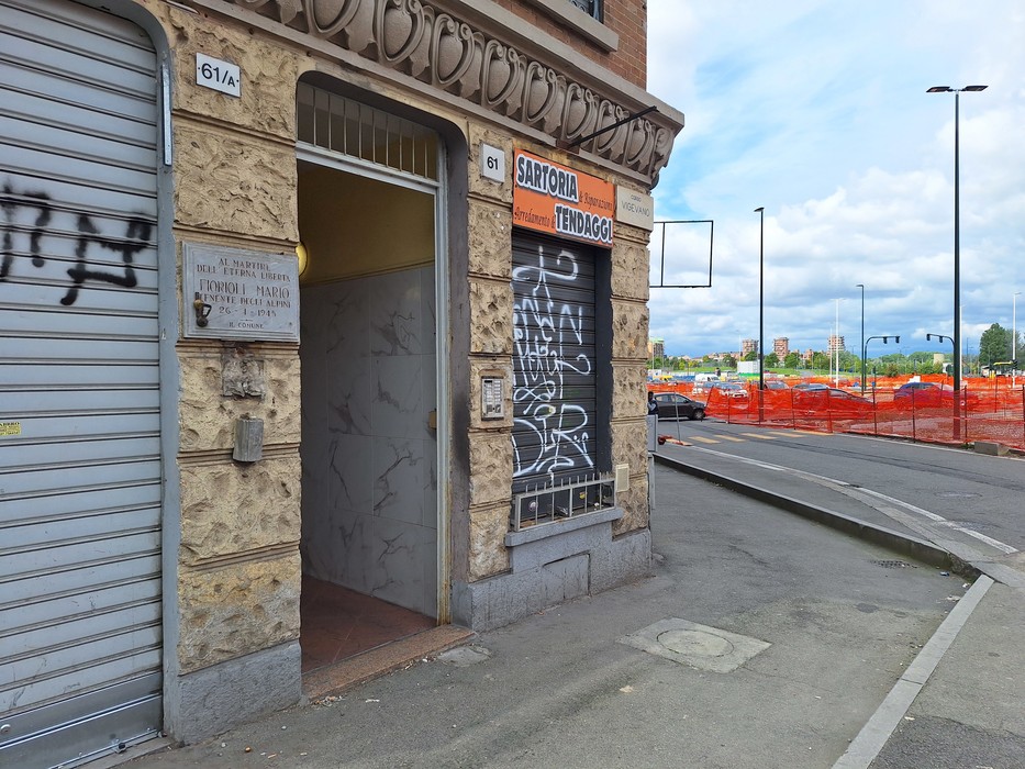 Ingresso di un edificio con scritte graffiti, accanto a un cantiere con recinzione arancione.
