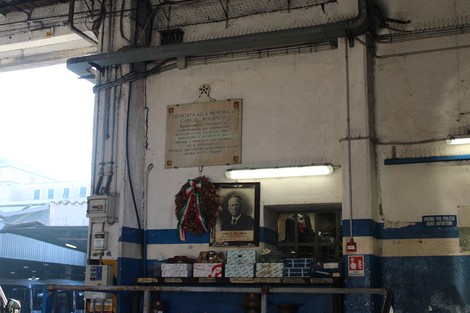 Lapide commemorativa di Rolando in un ambiente industriale, con decorazioni e fotografie storiche.