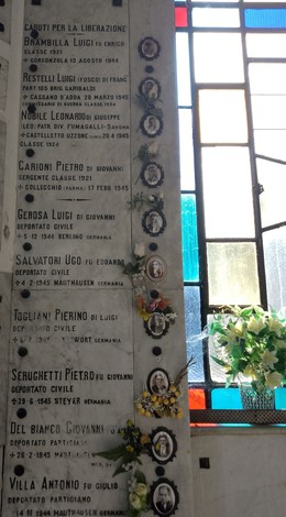 Lapide commemorativa con nomi di caduti per la libertà, decorata da fiori e fotografie in un contesto sobrio.