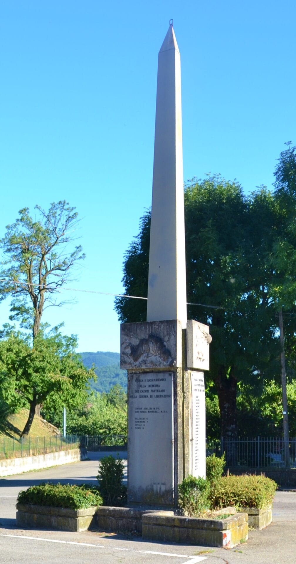 Obelisco commemorativo in onore dei caduti di Verica, circondato da un giardino e alberi, su uno sfondo blu.