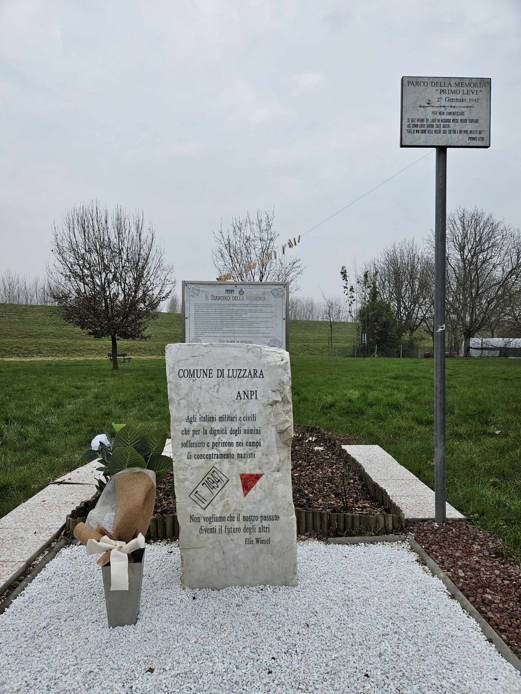 Cippo commemorativo dedicato agli internati e ai deportati, immerso nel verde, con decorazioni floreali.