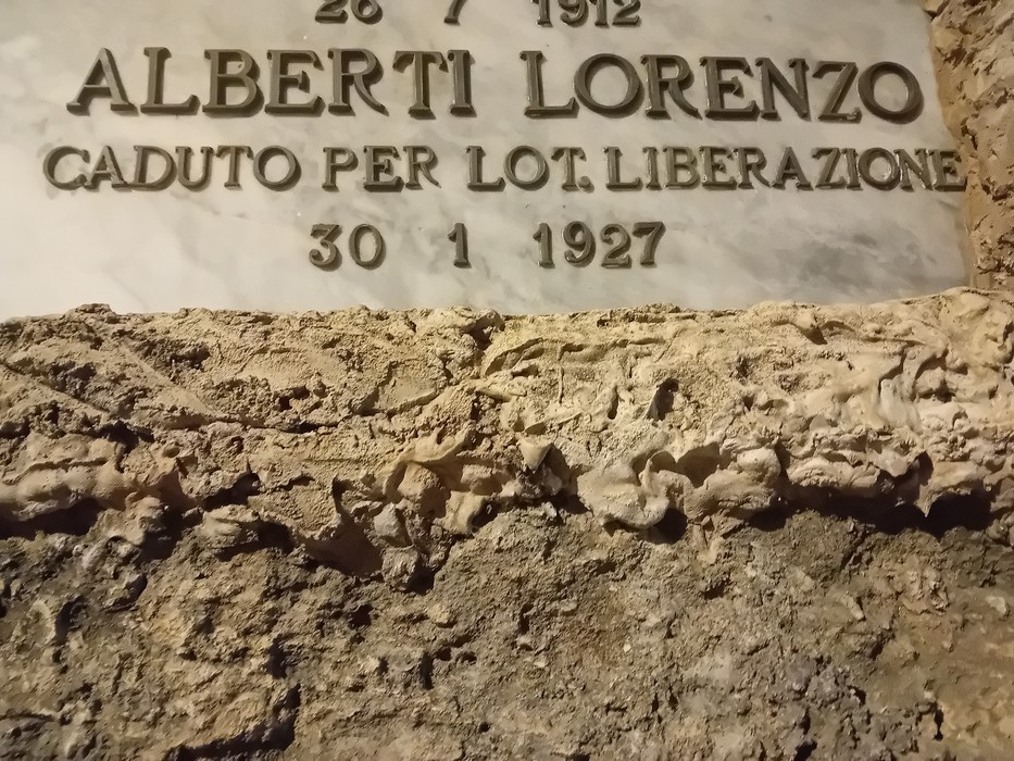 Lapide in marmo con iscrizione, fissata su una parete in pietra a vista.