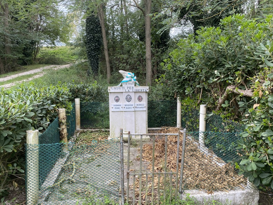Monumento commemorativo in un'area verde, circondato da vegetazione e rete, che onora Guerrini, Bedeschi e Morelli.