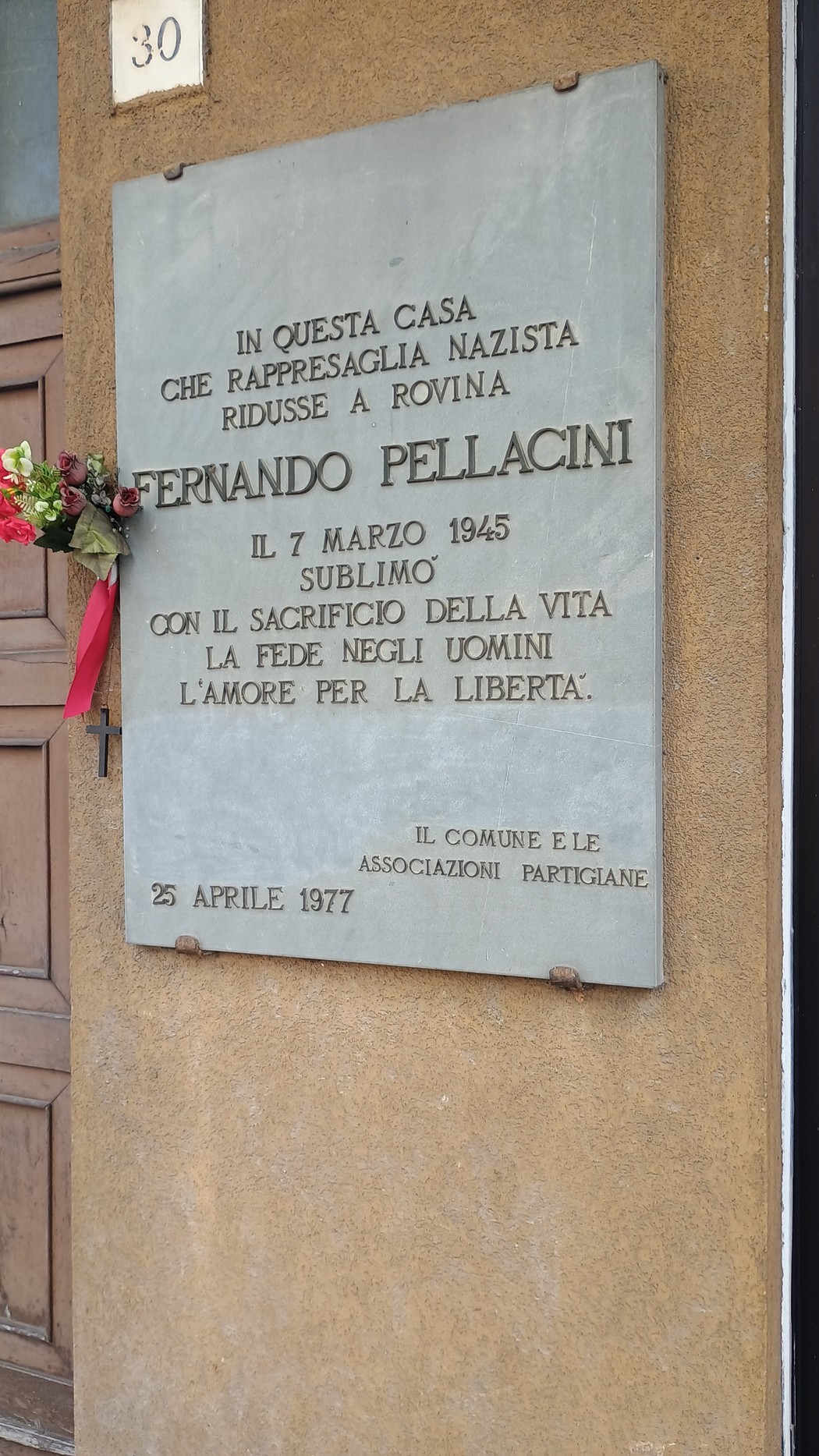 Lapide commemorativa dedicata a Fernando Pellacini, affissa su una parete esterna, con fiori decorativi.