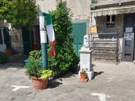 Cippo commemorativo con fiori e tricolore, situato in un contesto urbano.