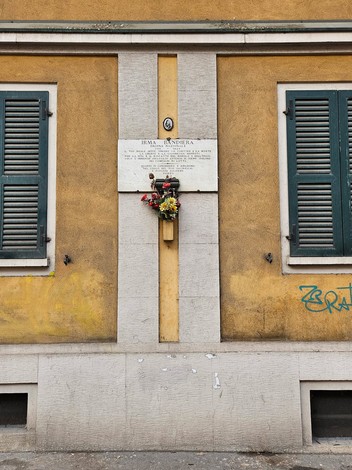 Lapide commemorativa su una parete gialla, con fiori e decorazioni, circondata da finestre verdi.