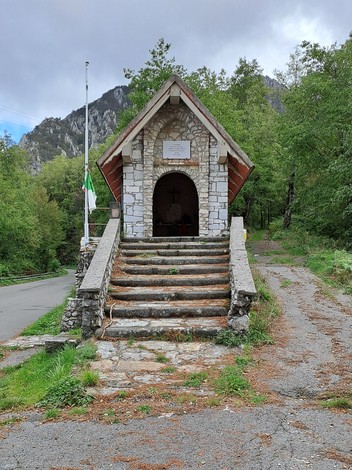 Sacrario in pietra con tetto spiovente e scalinata, arco d'ingresso con croce, targa sulla facciata, tra i boschi.