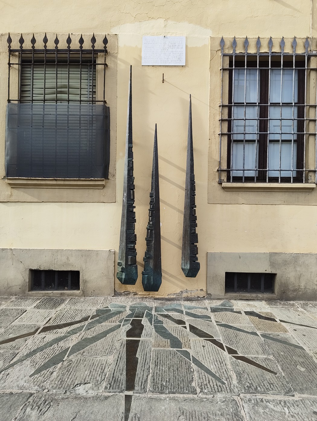Tre elementi metallici a forma di lama sulla facciata di un edificio, con lapide incisa in alto tra le finestre.