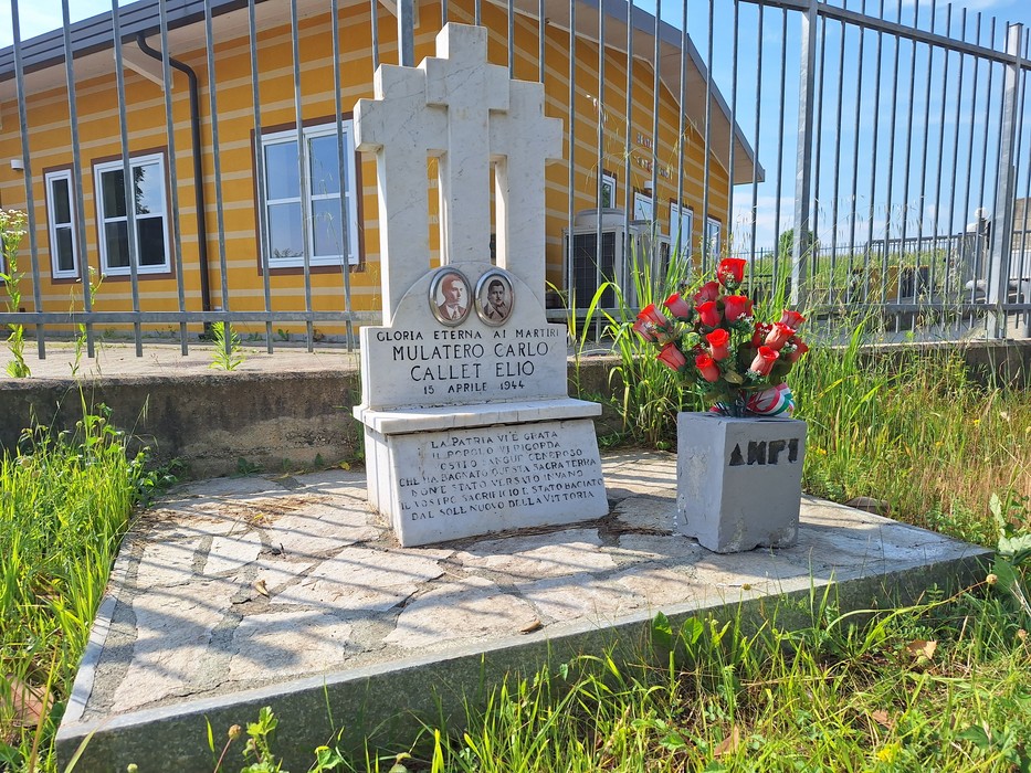 Cippo commemorativo in onore di Mulatero Carlo e Callet Elio, con fiori e decorazioni, immerso nel verde.