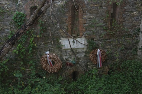 Lapide commemorativa con ghirlande e decorazioni, immersa nella vegetazione di un muro storico.