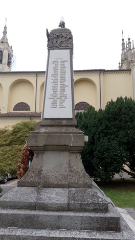 Monumento ai Caduti con lista di nomi, situato in un'area verde napoletana, sullo sfondo chiesa e alberi.
