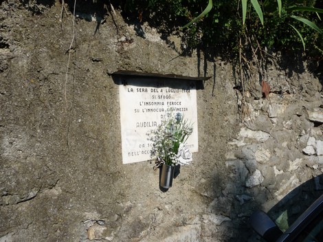 Lapide commemorativa su un muro, con fiori freschi e testo dedicato a Audilia Pellicioli.