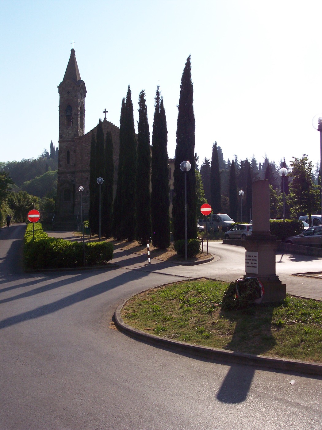 Cippo dei caduti con chiesa e filari di cipressi sullo sfondo in un paesaggio collinare.