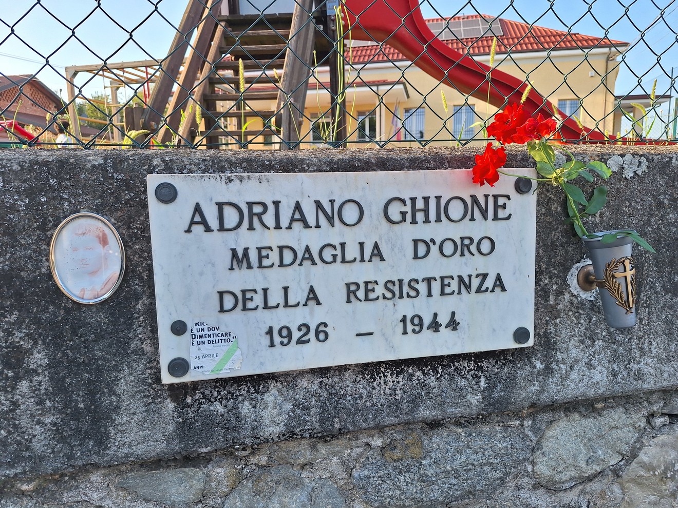Lapide in marmo con iscrizione, fissata su muro in pietra dietro una rete metallica, con fiori rossi accanto.