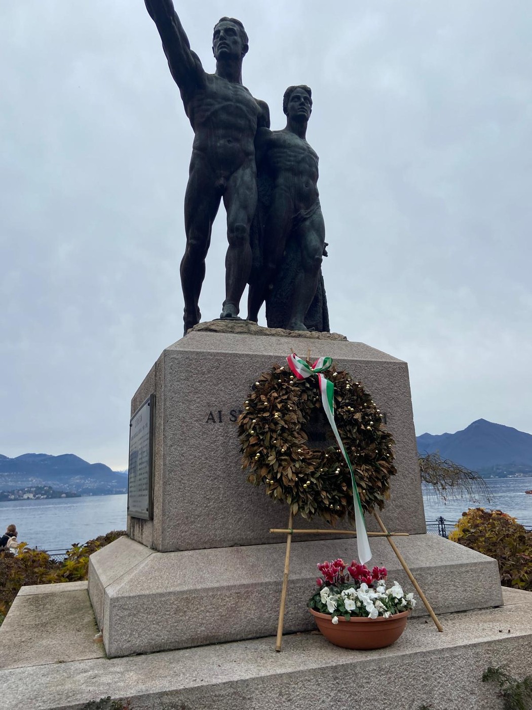 Monumento ai caduti con statue di soldati e una corona d'alloro ai piedi, vista lacustre sullo sfondo.
