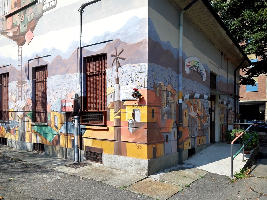 Murales colorato su una facciata di edificio, raffigurante un paesaggio urbano con case, montagne e un mulino.