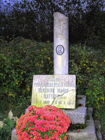 Cippo commemorativo dedicato a Mario Fanchini, circondato da fiori in un contesto naturale.