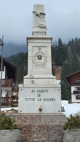 Monumento ai Caduti di Sappada, in marmo, circondato da alberi e abitazioni montane. Atmosfera di rispetto e memoria.