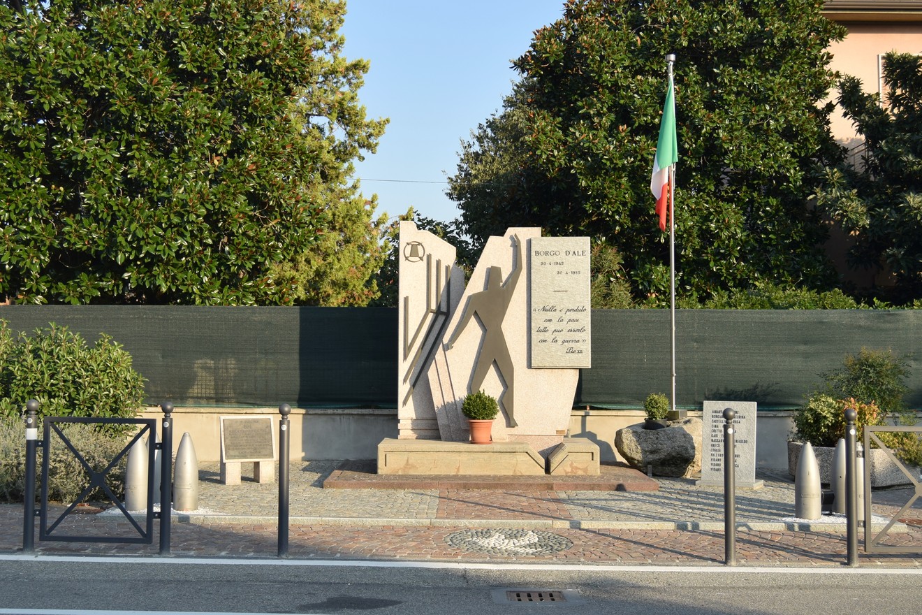 Monumento in pietra con sagoma di uomo a braccia alzate e iscrizione, in un'aiuola su strada con bandiera italiana.