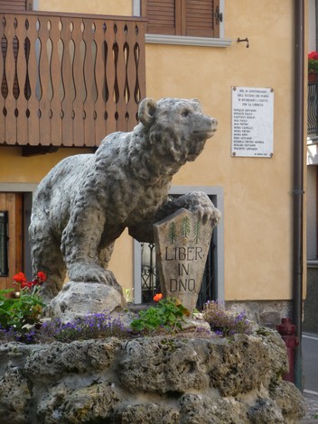 Scultura di un orso con una lapide che celebra i Dodici Caduti per la Libertà, decorata con fiori colorati.