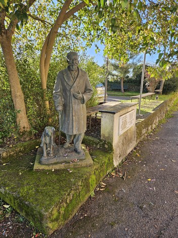 Monumento a Benedicenti, statua bronzea con uomo in cappotto e cane, immersi nel verde di un parco.