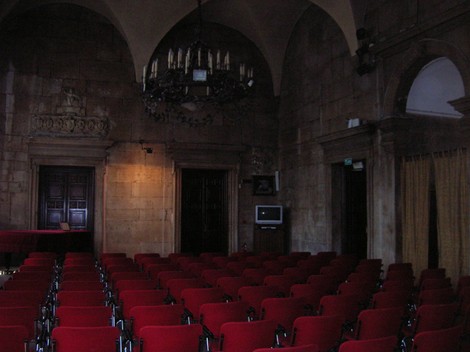 Interno di una sala con sedie rosse disposte ordinatamente, illuminata da un lampadario elegante e decorazioni murali.