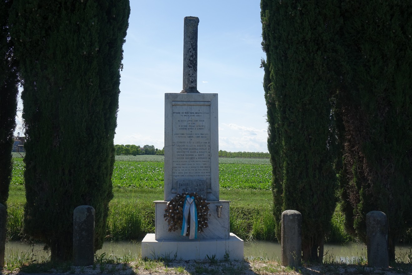Monumento commemorativo dedicato a Gennaro con una corona di fiori, circondato da alberi e campo verde sullo sfondo.
