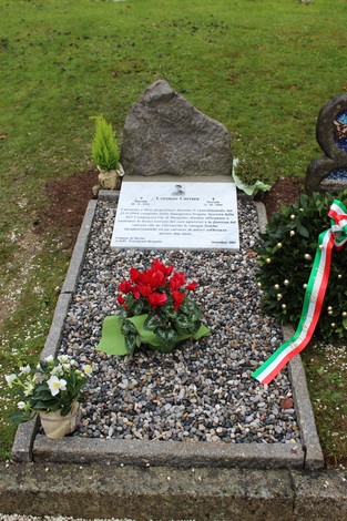 Cippo commemorativo a Carrara, adornato con fiori e decorazioni, in un contesto verde e sereno.