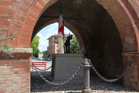 Monumento ai Caduti di Piumazzo, con statue e tricolore, sotto un arco di mattoni in un contesto urbano.