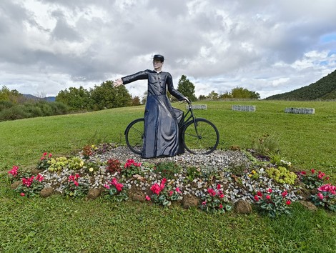 Statua di un angelo su una bicicletta circondata da fiori, in un contesto naturale con cielo nuvoloso.